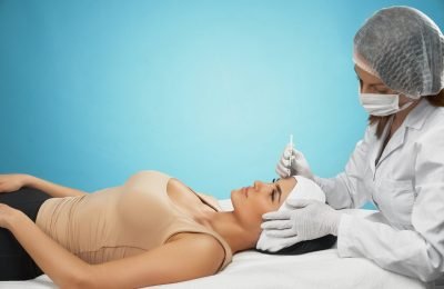 Side view of injection in female forehead. Cosmetologist in doctors coat using syringe with special liquid, brunette patient lying, isolated on blue. Concept of cosmetology, beauty.